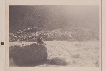 It appears to be Bessie Hyde sitting next to a rapid which flows from left to right.  Print owned by Mrs. William Haley and probably one of those made from the films found in the snow