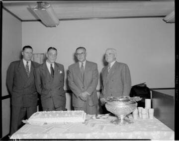 Four men standing in front of a large sheet cake and punch bowl